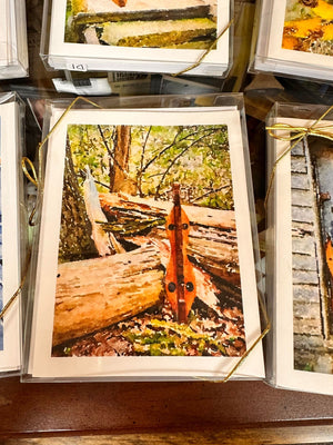 A framed painting by Ellen Pratt shows a wooden string instrument leaning against tree logs in a forest setting. The frame is tied with a golden ribbon, evocative of her Dulcimer Greeting Card Sets by Ellen Pratt.