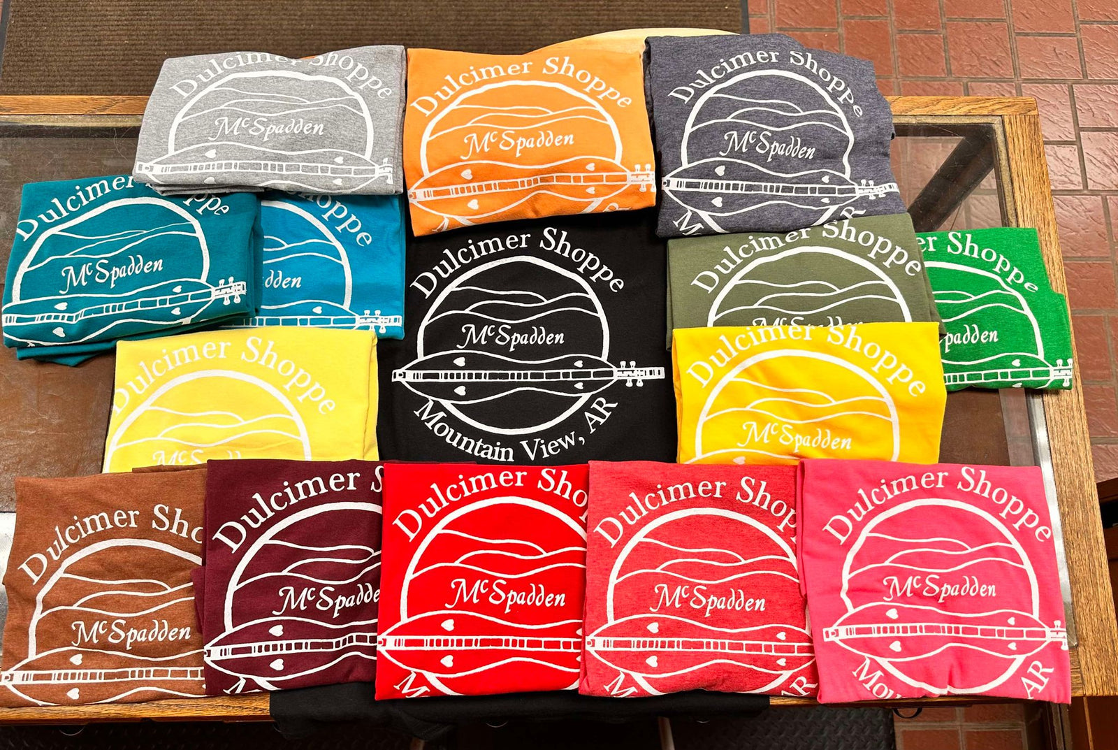 A display of McSpadden Dulcimer Shoppe Logo Shirts from Mountain View, AR, showcases a variety of colors and sizes, featuring blue, orange, yellow, green, red, and gray options.
