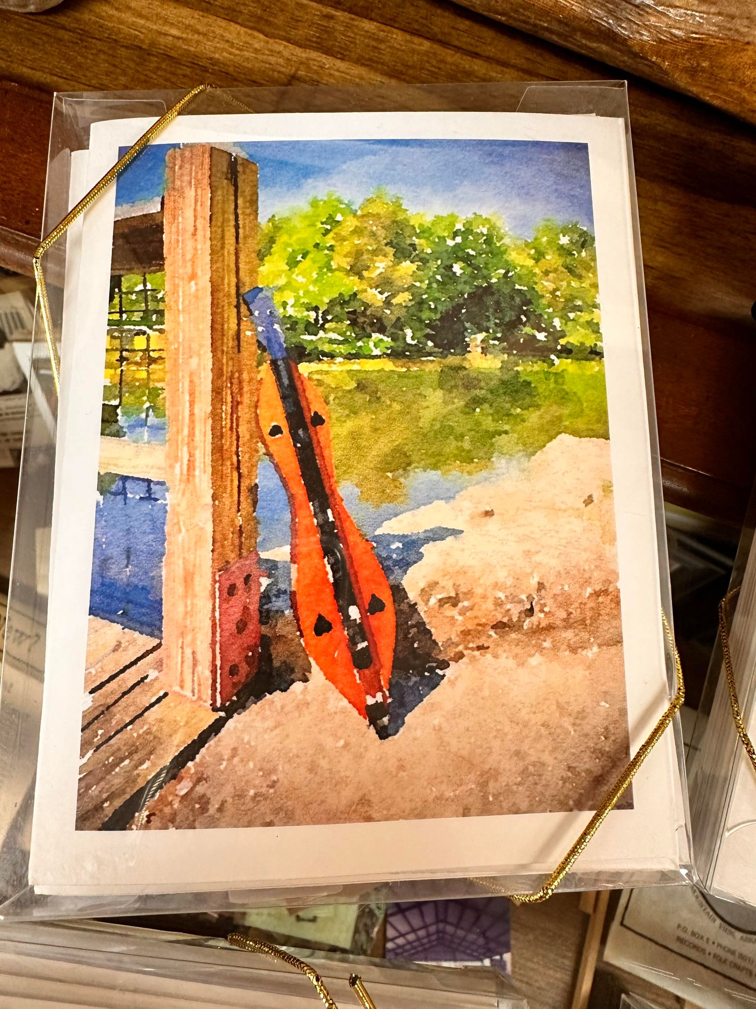 A painting of a lakeside scene with a musical instrument, possibly a dulcimer, leaning against a wooden post on a dock. Trees and water are in the background, reminiscent of Dulcimer Greeting Card Sets by Ellen Pratt.