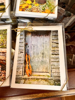 A stack of Dulcimer Greeting Card Sets by Ellen Pratt features a watercolor painting of a weathered door with a yellow violin leaning against it. The cards, tied with a gold ribbon, capture an artistic charm perfect for any occasion.