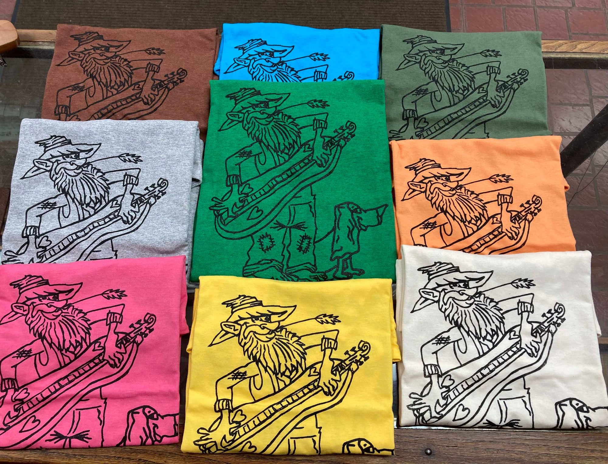 Nine "Shirt with Hillbilly" short-sleeved shirts in various colors, each featuring a black line drawing of a hillbilly character with a guitar and a dog.