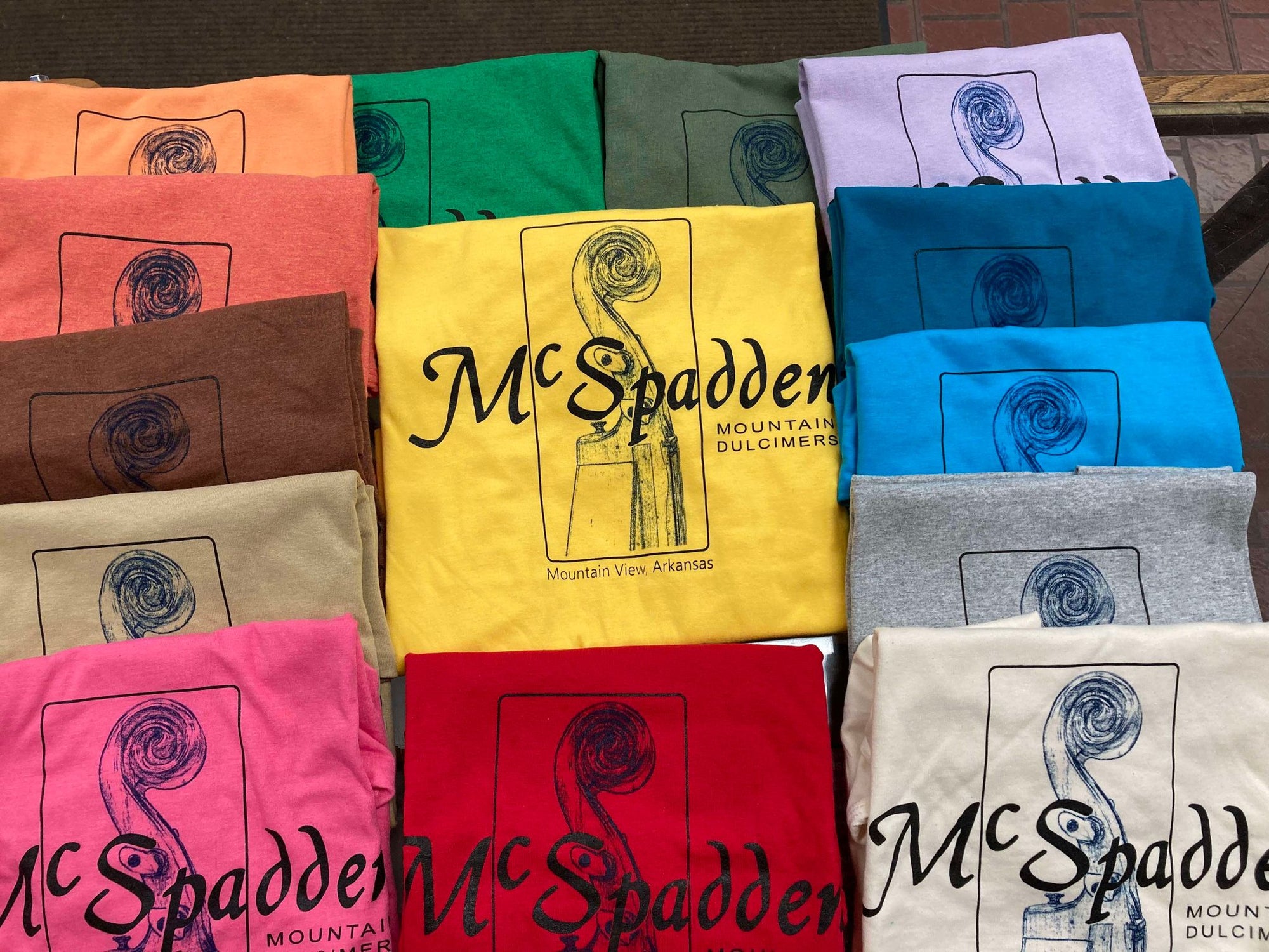 Vibrant and assorted short-sleeved "Shirt with Scroll Head McSpadden" featuring the text "McSpadden Mountain Dulcimers" and a dulcimer graphic, available in multiple colors and sizes.