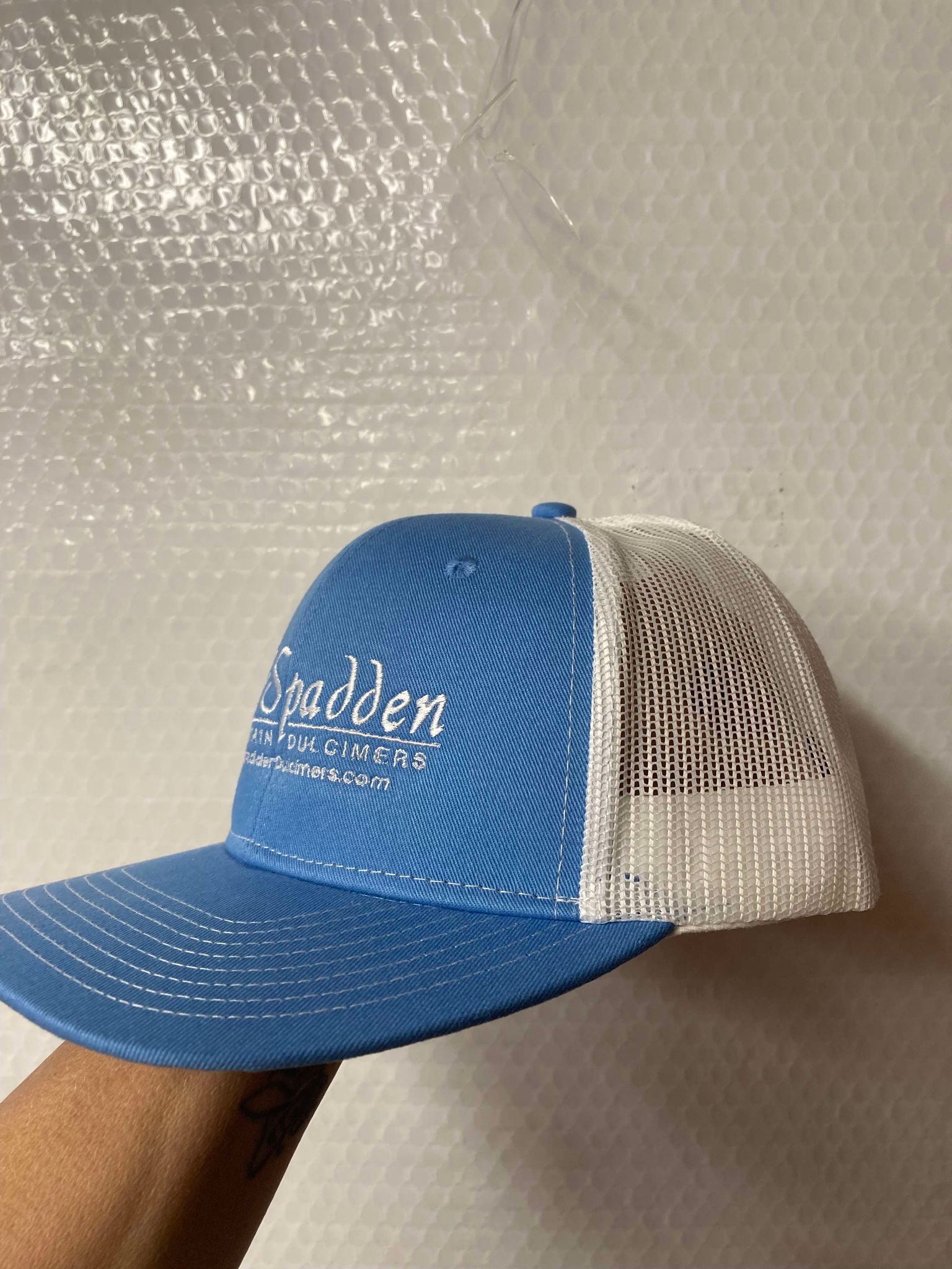 Introducing the McSpadden Dulcimer Trucker Hat: a stylish blue and white trucker hat featuring a high front panel and the text "Spadden Mountain Dulcimers" elegantly embroidered on the front.