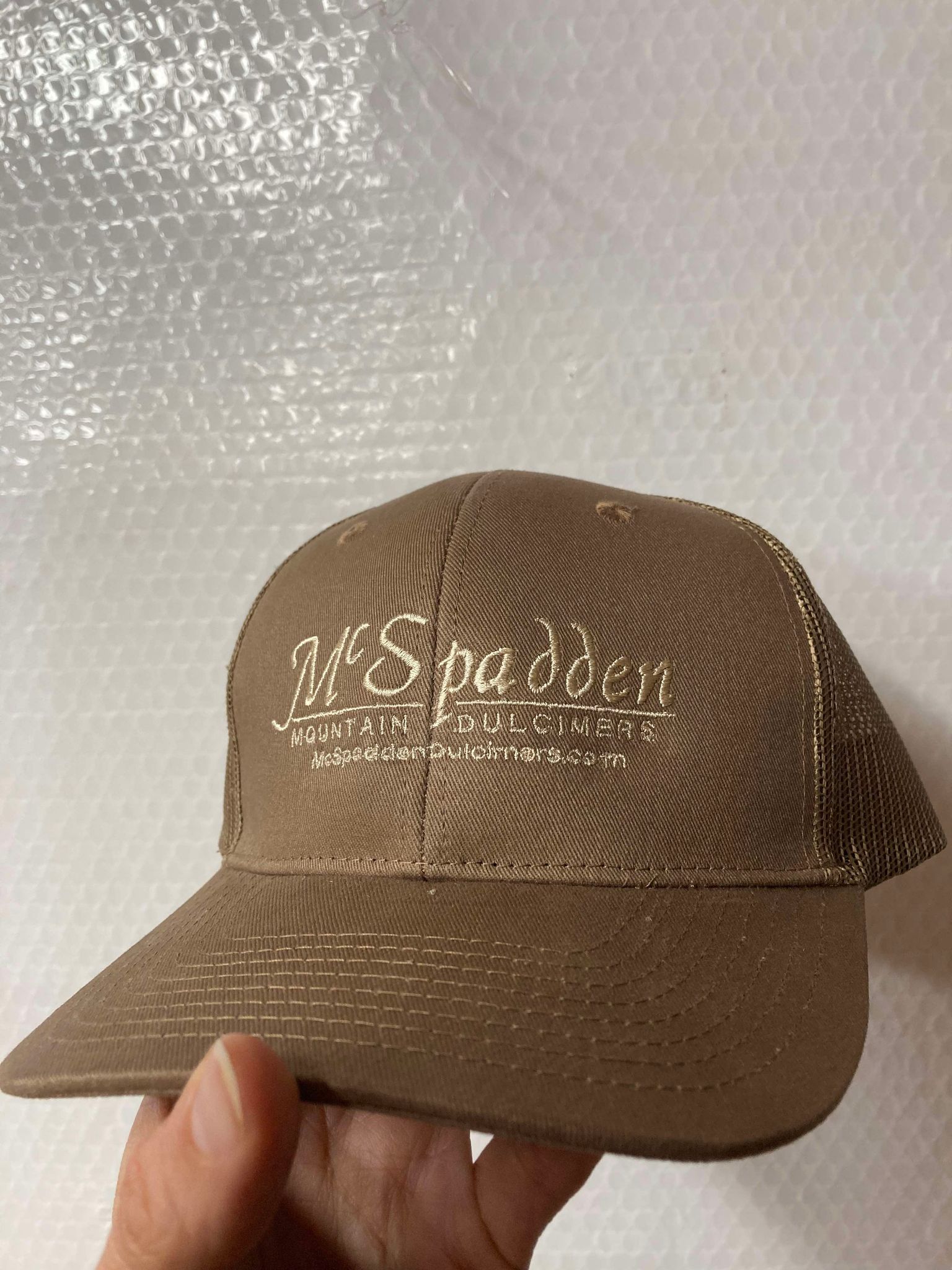 A hand holds the McSpadden Dulcimer Trucker Hat, featuring a brown design and the "McSpadden Mountain Dulcimers" logo embroidered on the front, against a white textured background. This cool-vibe accessory is your perfect road trip buddy.