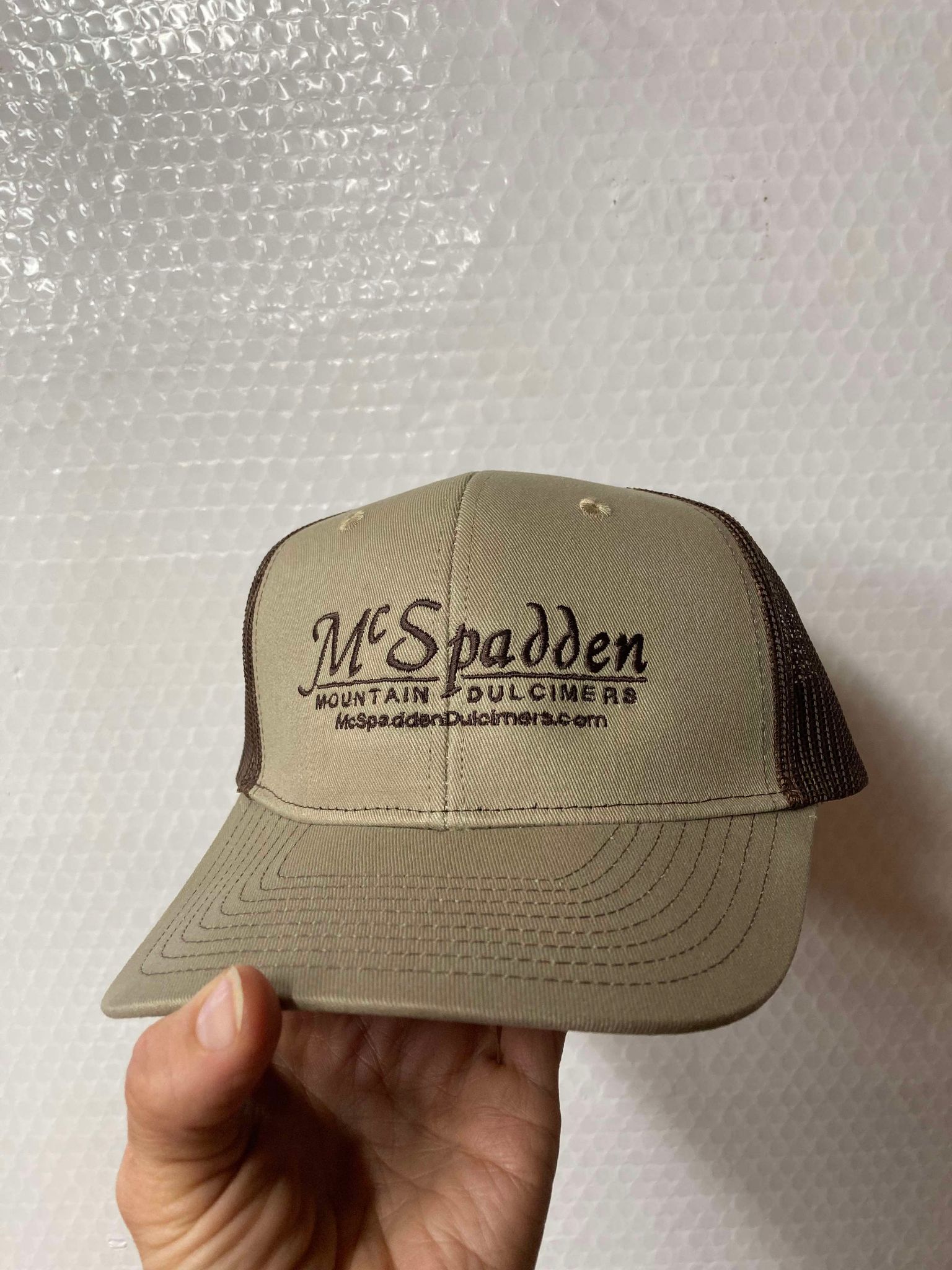 A hand holds a McSpadden Dulcimer Trucker Hat featuring a high beige and brown front panel embroidered with the "McSpadden Mountain Dulcimers" logo and the website "McSpaddenDulcimers.com." The mesh back guarantees excellent breathability.