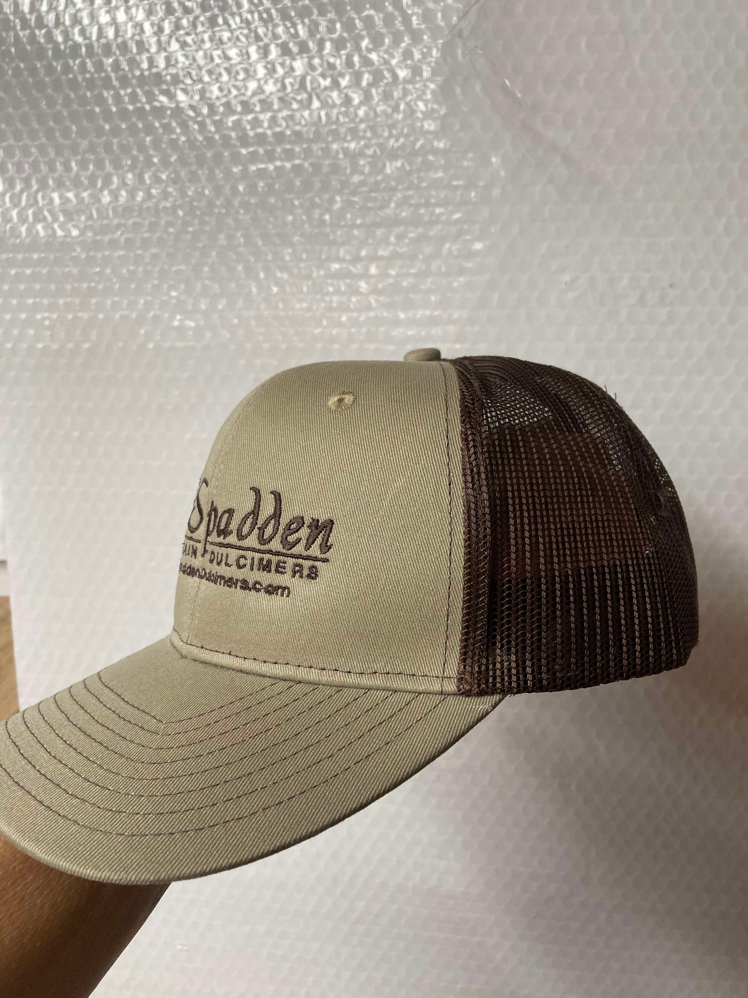 A McSpadden Dulcimer Trucker Hat with a beige and brown mesh design, featuring the embroidered text "Spadden Custom Builders" on the front, held in one hand against a bubble wrap background—your perfect road trip buddy.