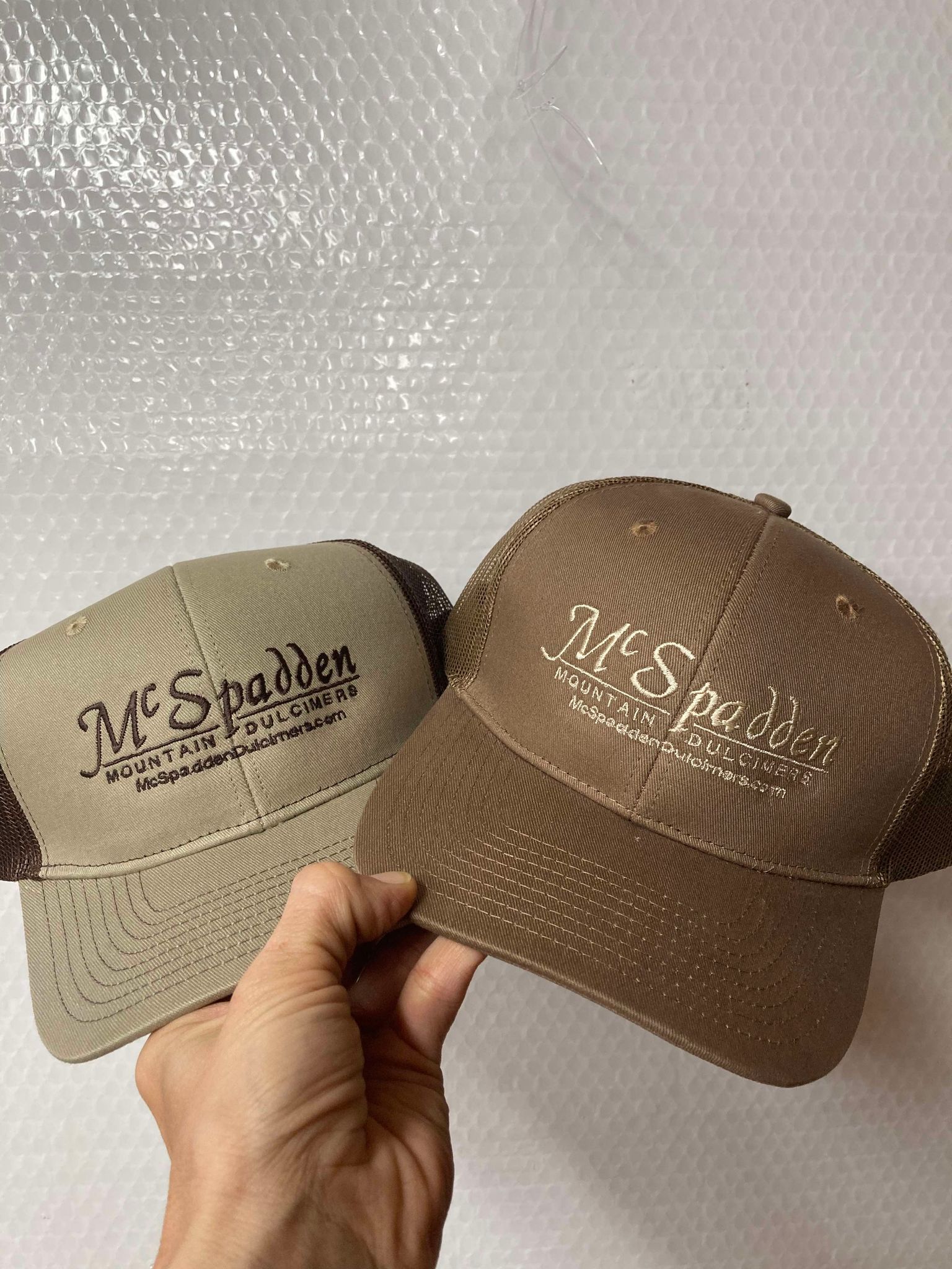 Two McSpadden Dulcimer Trucker Hats, perfect for road trip buddies, are held side by side. Both hats have "McSpadden Mountain Dulcimers" printed on the front, showcasing their cool vibe with one in light tan and the other in darker brown.