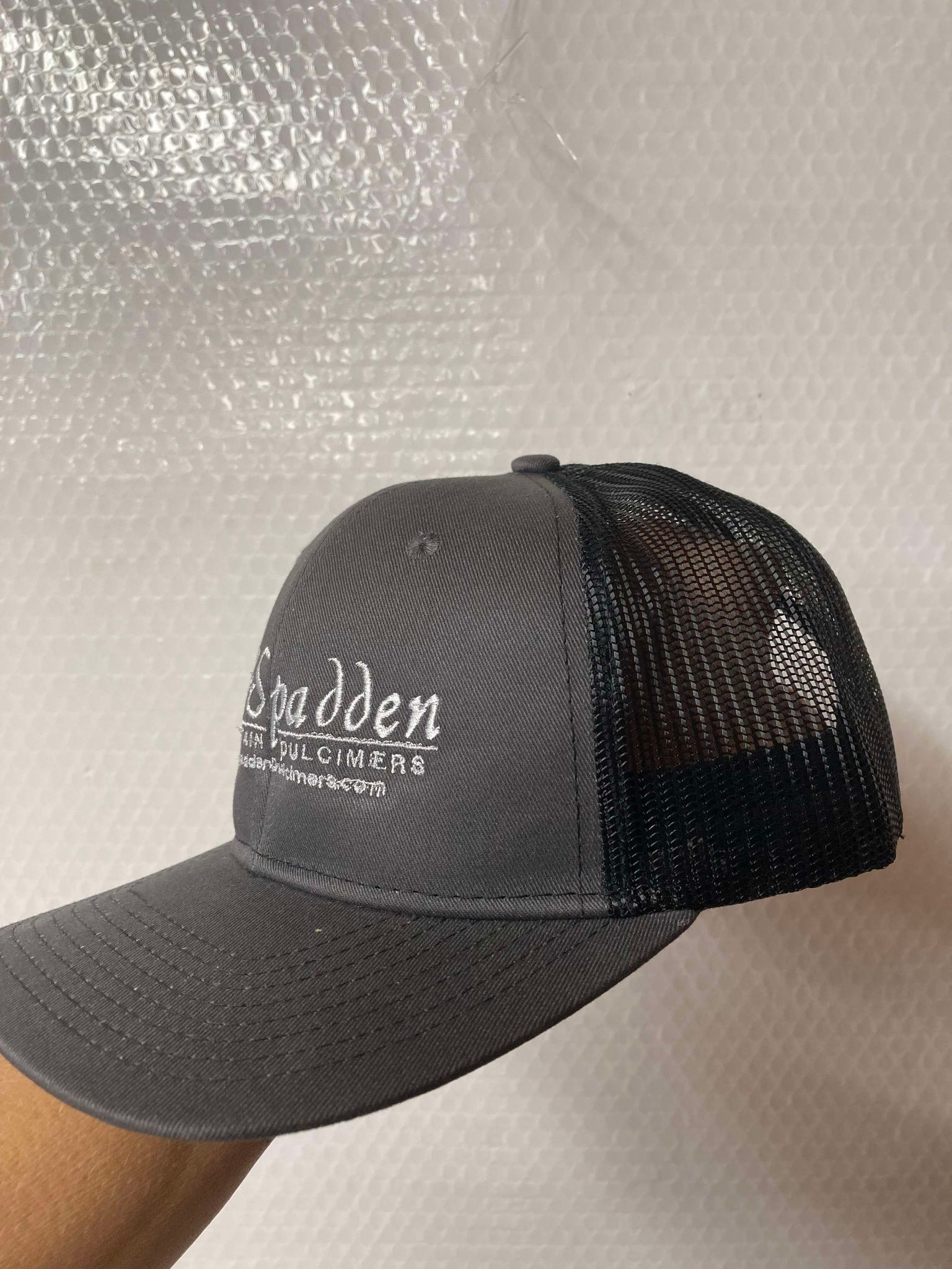 Close-up of a McSpadden Dulcimer Trucker Hat showcasing its high-front gray panel and black mesh back, adorned with a white embroidered "Spadden Dulcimers" logo along with additional text and the website at the bottom.