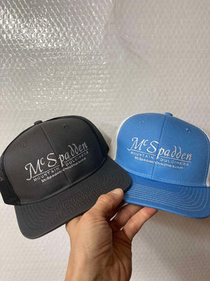 A hand is holding two McSpadden Dulcimer Trucker Hats, both featuring "McSpadden Mountain Dulcimers" logos and a high front panel design; one hat is black and gray, while the other is blue and white with a mesh back.