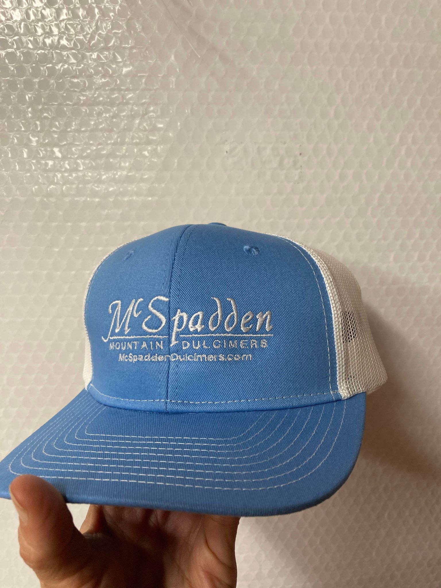 A hand holds the McSpadden Dulcimer Trucker Hat, showcasing its blue and white design with "McSpadden Mountain Dulcimers" and "McSpaddenDulcimers.com" embroidered on the high front panel. The mesh back ensures breathability, making it ideal for long days outdoors.