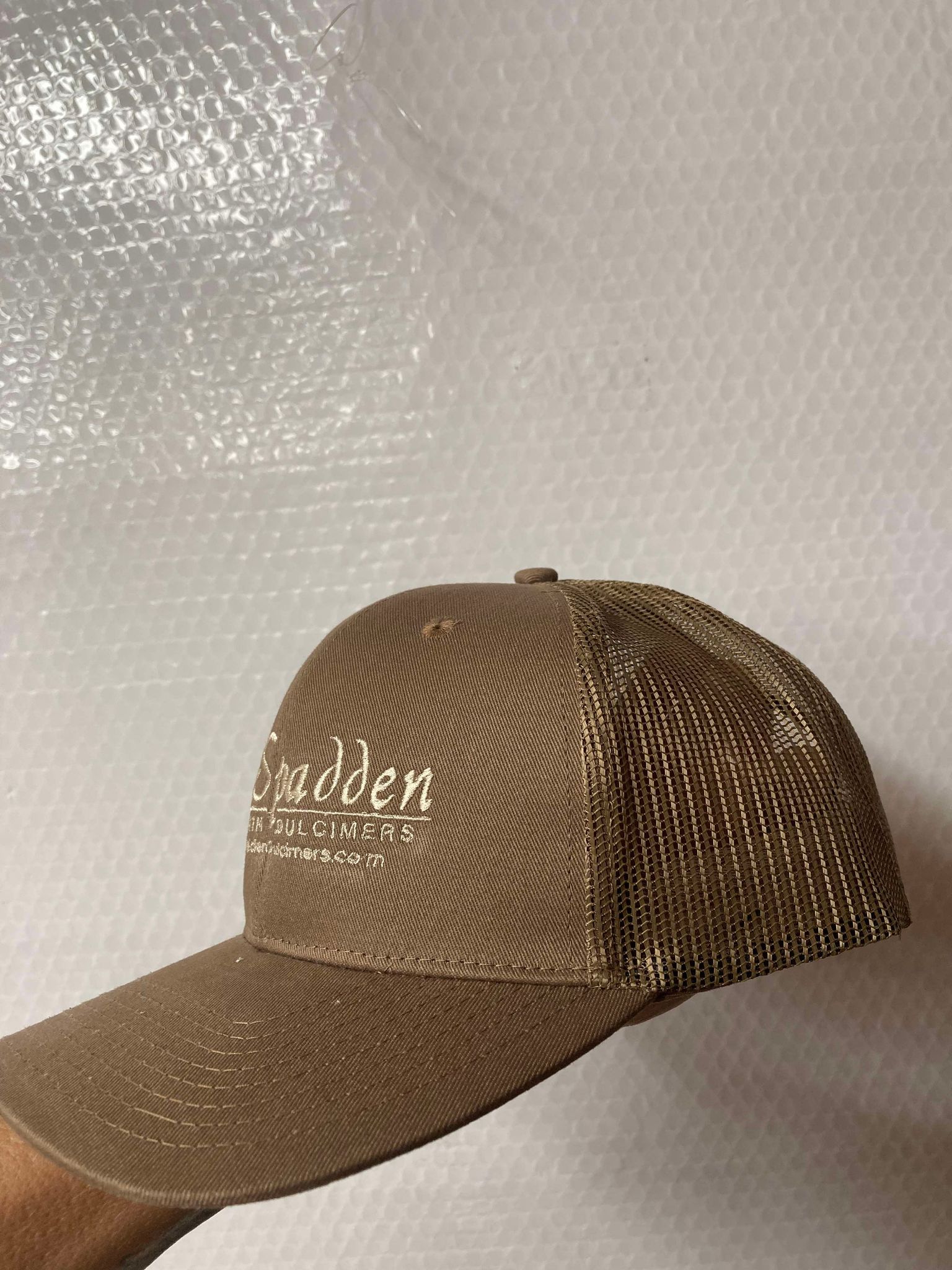 A McSpadden Dulcimer Trucker Hat featuring a brown mesh-back, high front panel, and the word "Spadden" embroidered on the front is held up against a textured white background.