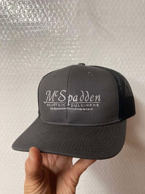 A hand holding a McSpadden Dulcimer Trucker Hat, which has "McSpadden Mountain Dulcimers" embroidered on the high front panel and the website text below. The gray and black hat features a stylish mesh back for breathability.