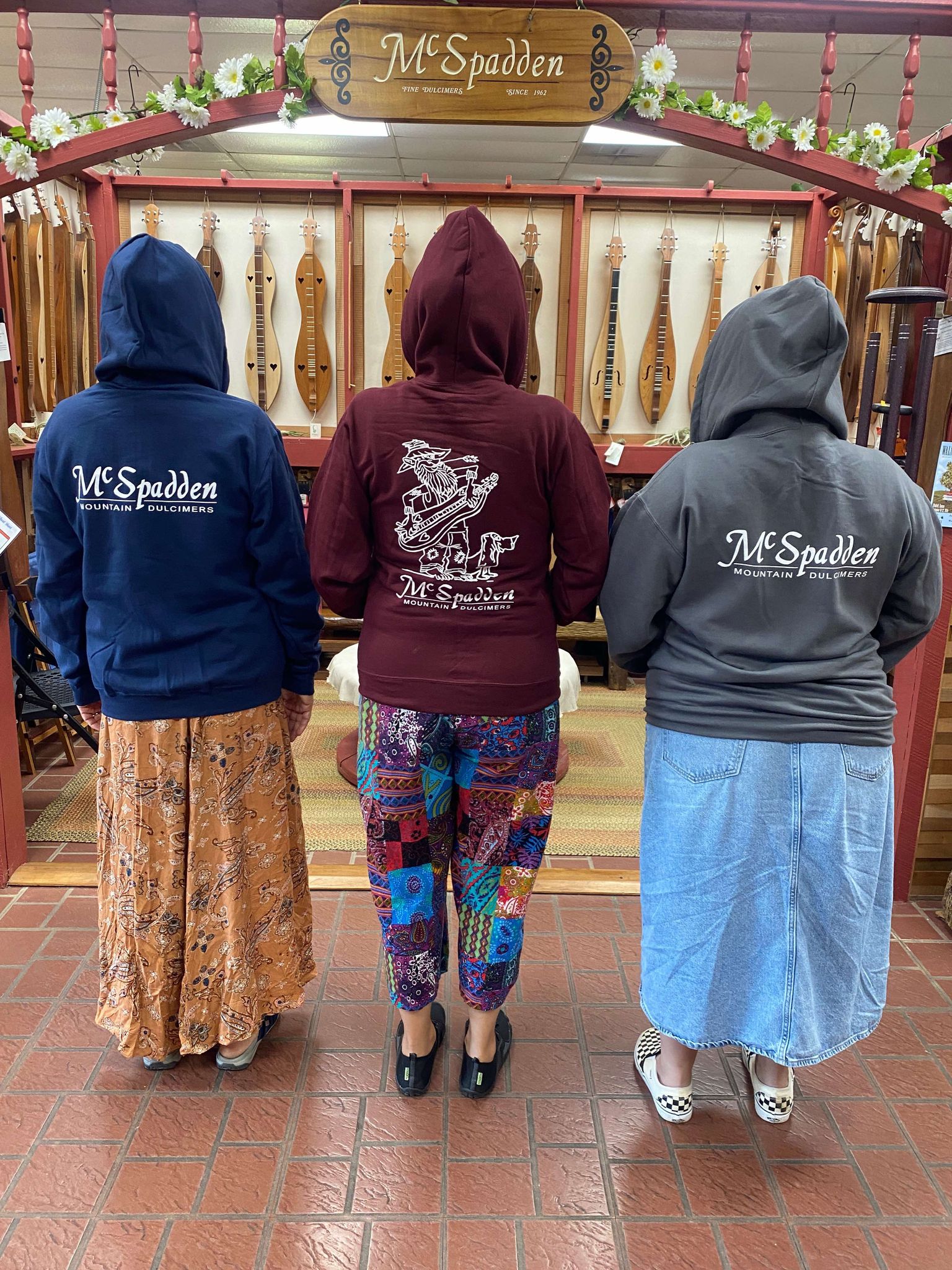 Three people stand with their backs to the camera, sporting Dulcimer Shoppe Hoodies that feature "McSpadden Mountain Dulcimers" logos. They are in a shop showcasing high-quality dulcimers in the background.