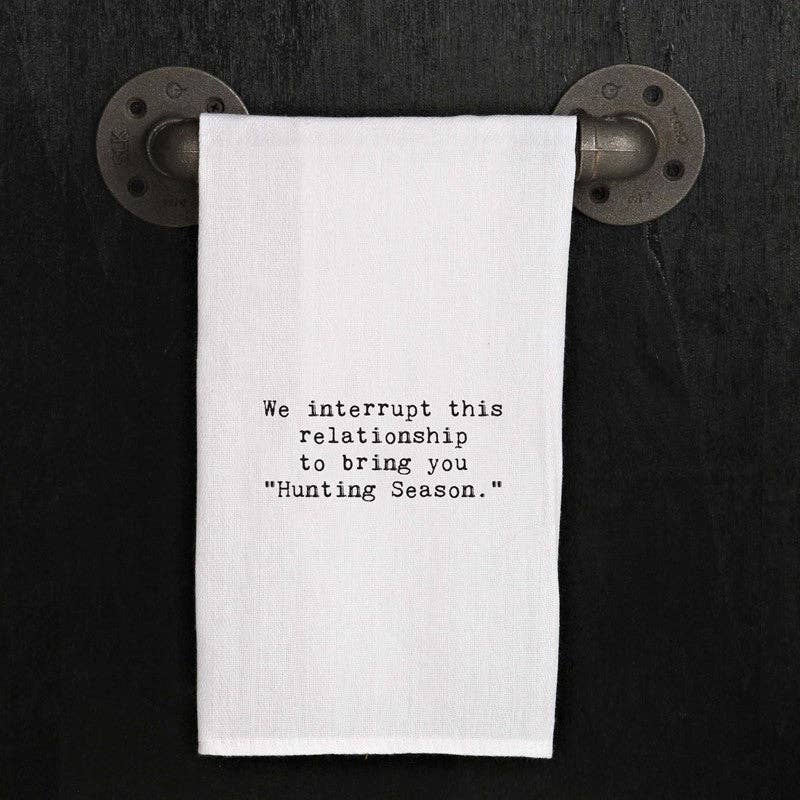 The "We interrupt this relationship to bring you 'Hunting Season'" white hand towel is crafted from 100% cotton and hangs on a metal pipe-style towel holder against a dark background. This versatile kitchen towel makes an ideal handmade hostess gift.