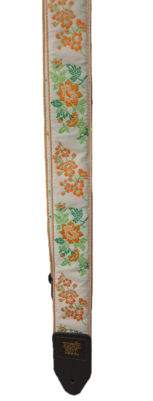 The Alpine Meadow Guitar Strap is cream-colored with orange and green floral Jacquard embroidery, finished with a black leather end labeled "Ernie Ball" and model number EB4617.