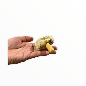 A small handmade wooden frog toy, crafted in Thailand with quality craftsmanship, is held in a person's hand. The toy features a cylindrical protrusion from its mouth.