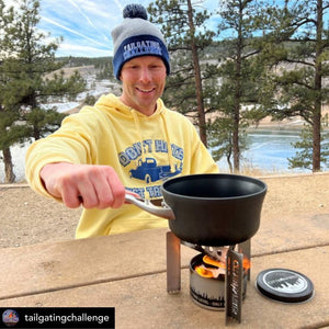 Man in yellow hoodie and blue beanie cooking on a Portable Camp Stove / Stand Combo outdoors, with a scenic lake and trees in the background.