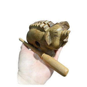 A hand holds the Wooden Musical Pig Percussion Instrument, handcrafted in Thailand with a ridged back and a wooden stick for playing.
