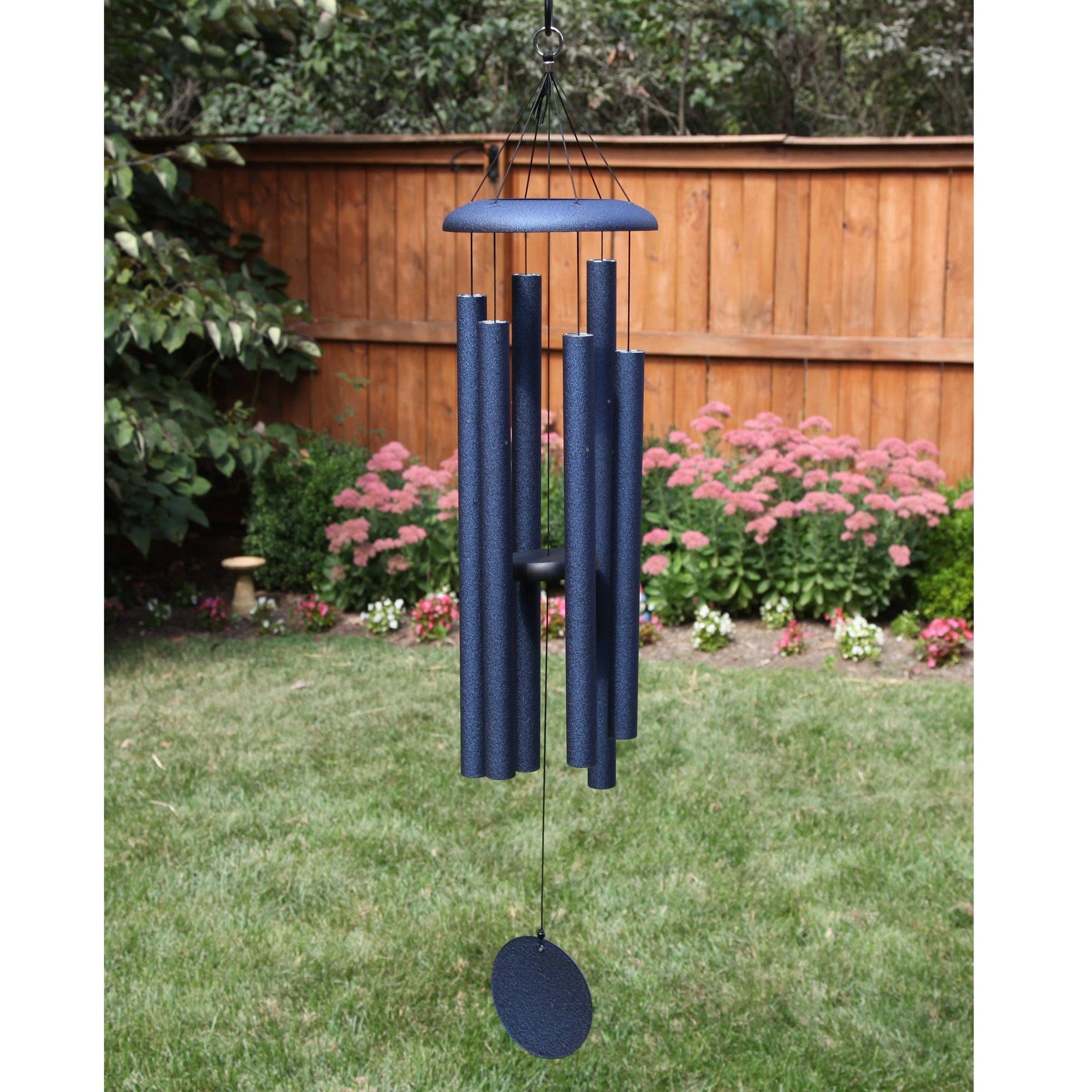 A Corinthian Bells® 44-inch Windchime with flair hangs in a backyard with a wooden fence, green lawn, and pink flowering plants.