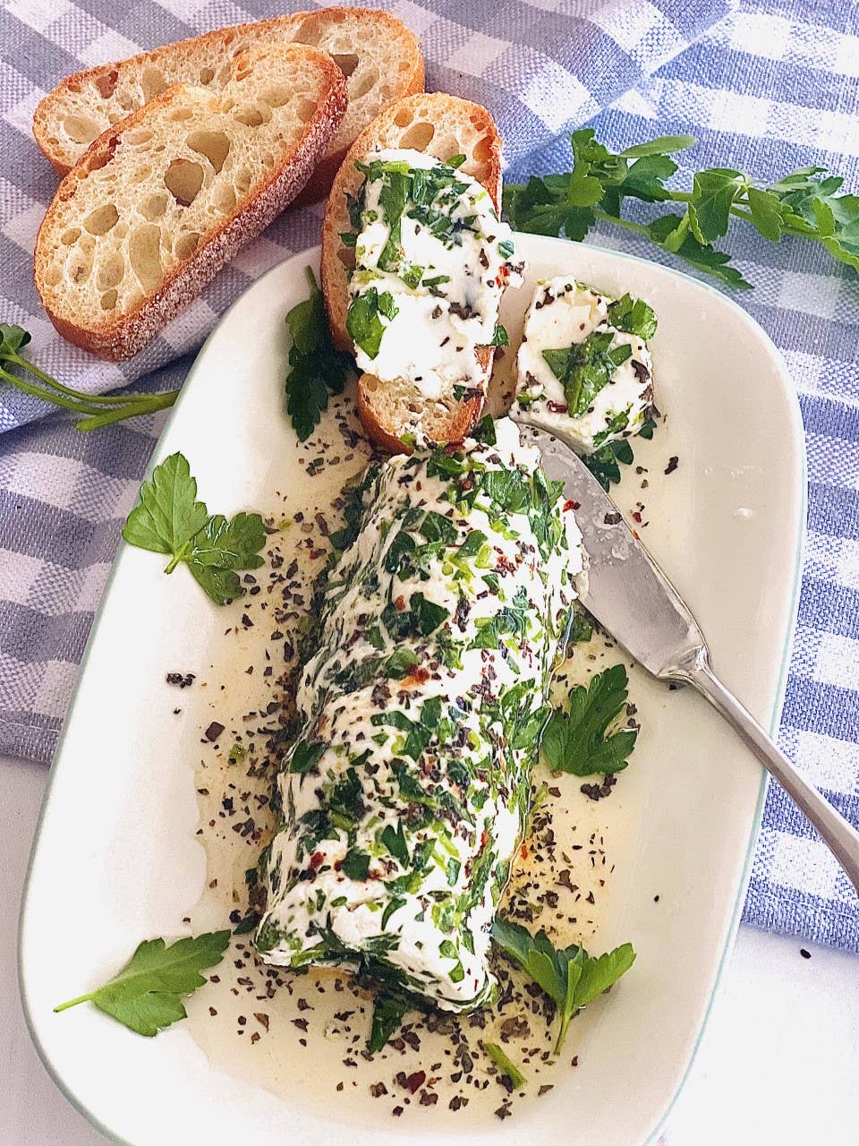 A log of goat cheese coated in herbs and pepper, topped with a creamy white sauce infused with Carmie's Pesto Dipping Oil Seasoning, served with slices of toasted baguette on a white plate. A blue and white checkered cloth is in the background.