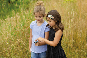 Two young girls examining an Outdoor Discovery Backyard Exploration Clip-On Compass together in a field of tall grass. One girl is wearing a black dress, and the other a blue t-shirt.