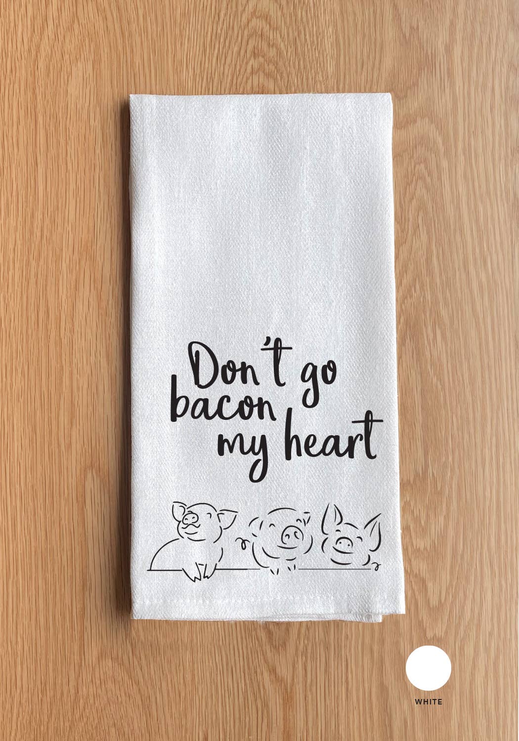 A "Don't go Bacon my heart (pig icon)" kitchen towel, showcasing the phrase along with three charming pig icons at the bottom, elegantly displayed on a wooden surface.