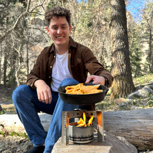 Young man smiling, sitting outdoors by a Portable Camp Stove / Stand Combo with a pan of food, surrounded by trees.