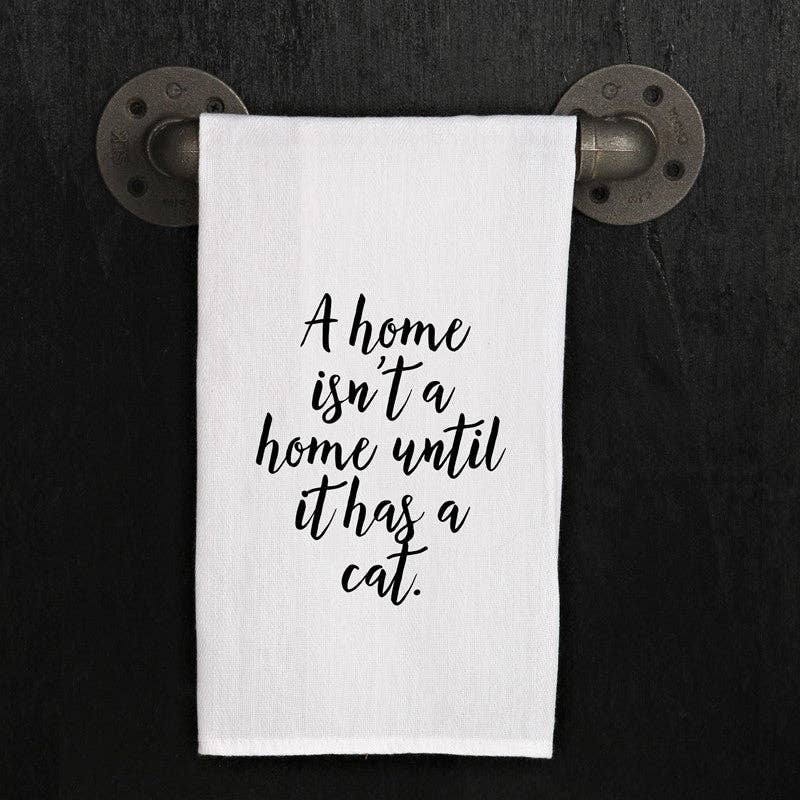 A white, 100% cotton towel hangs on a metal pipe holder, featuring the text "A home isn't a home until it has a cat." This versatile kitchen towel, named "A home isn't a home until it has a cat," makes for the perfect handmade hostess gift.