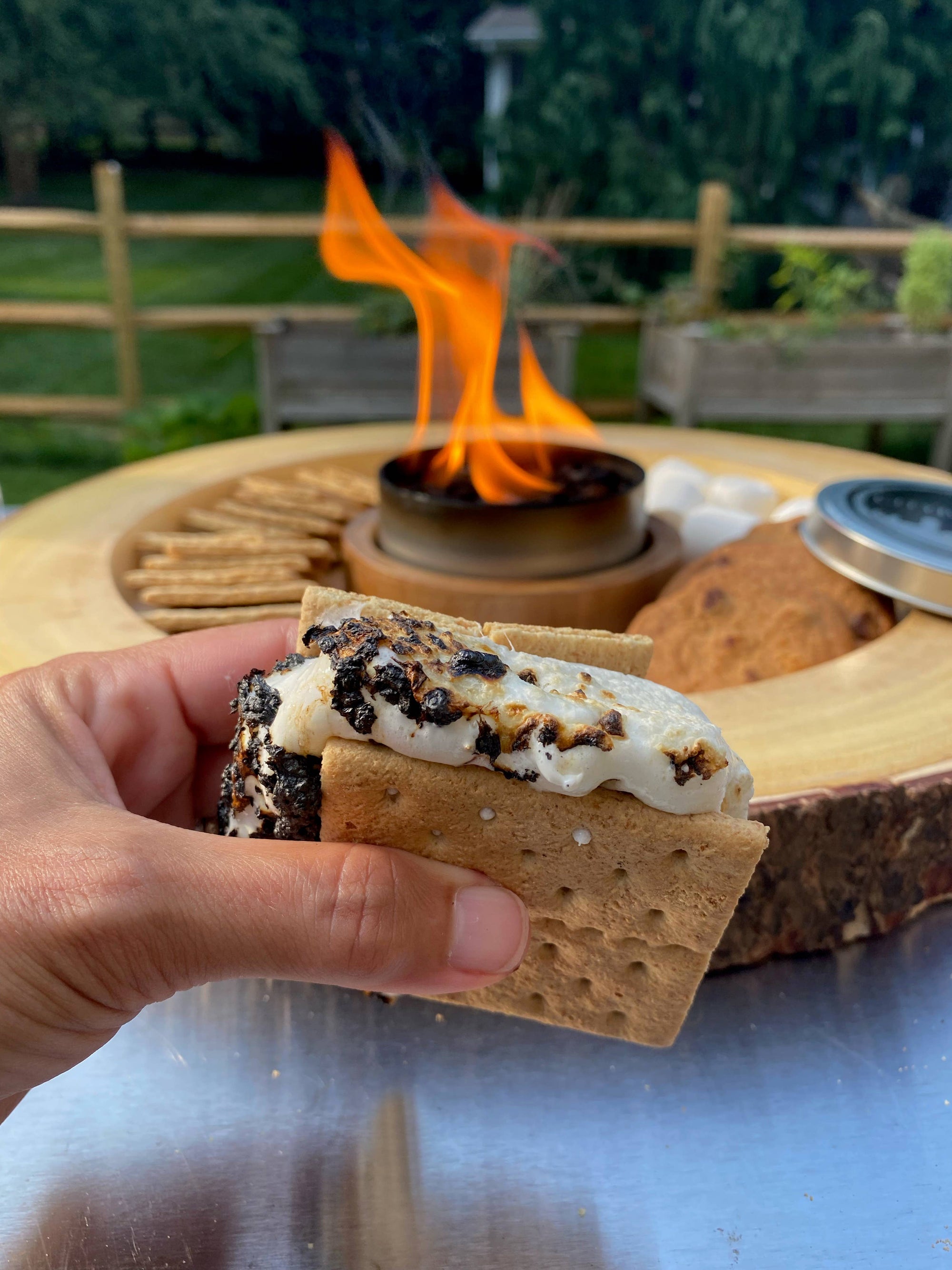 A person holding a s'more with a marshmallow and chocolate between graham crackers, with a City Bonfires® by Coleman® in the background.