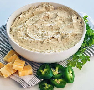 The Jalapeno Cheddar Dip Mix sits on a striped cloth, surrounded by sliced jalapeños, chiles, cheese cubes, and parsley sprigs—ideal for sharing at your next holiday gathering!.