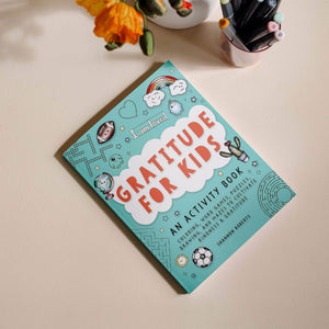 A copy of "Gratitude For Kids (Mindfulness Activity Book for Kids)" rests on a table beside orange flowers and a cup of pens—ideal for anyone seeking mindfulness activity books for kids.