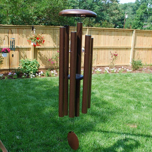 A Corinthian Bells® 60-inch Windchime in brown hangs in a grassy backyard with a wooden fence, garden, and trees in the background, filling the air with melodious tones reminiscent of a major 7th chord.