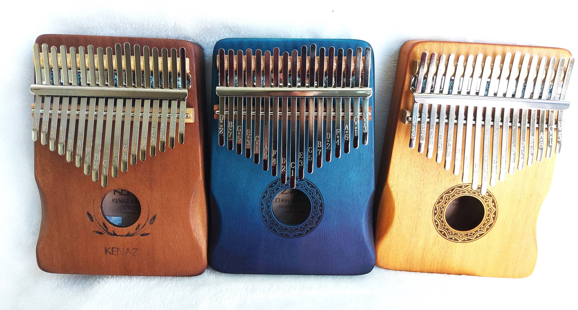 Three Kalimba Musical Instruments with varying wood tones (light, blue, and medium brown) displayed on a white surface, each with metal tines and decorative sound holes.