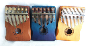 Three Kalimba Musical Instruments with varying wood tones (light, blue, and medium brown) displayed on a white surface, each with metal tines and decorative sound holes.