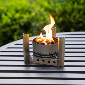 A Portable Camp Stove / Stand Combo with a visible flame, placed on a striped outdoor table, surrounded by green foliage, doubles as a collapsible camp stove.