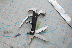 A versatile Black Wood Multi Hammer Tool with various tools unfolded, lying next to a metal ruler on a work surface with ink marks and cuts.