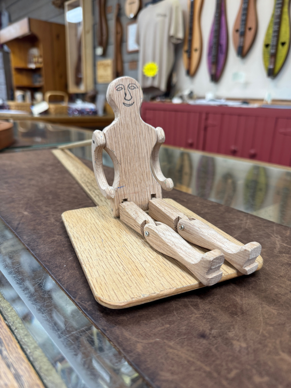 The Dancing Man Toy, a wooden figurine with jointed limbs and a smiling face on a flat wood base, is featured in Thomas Dunn’s shop among musical instruments and wooden crafts.