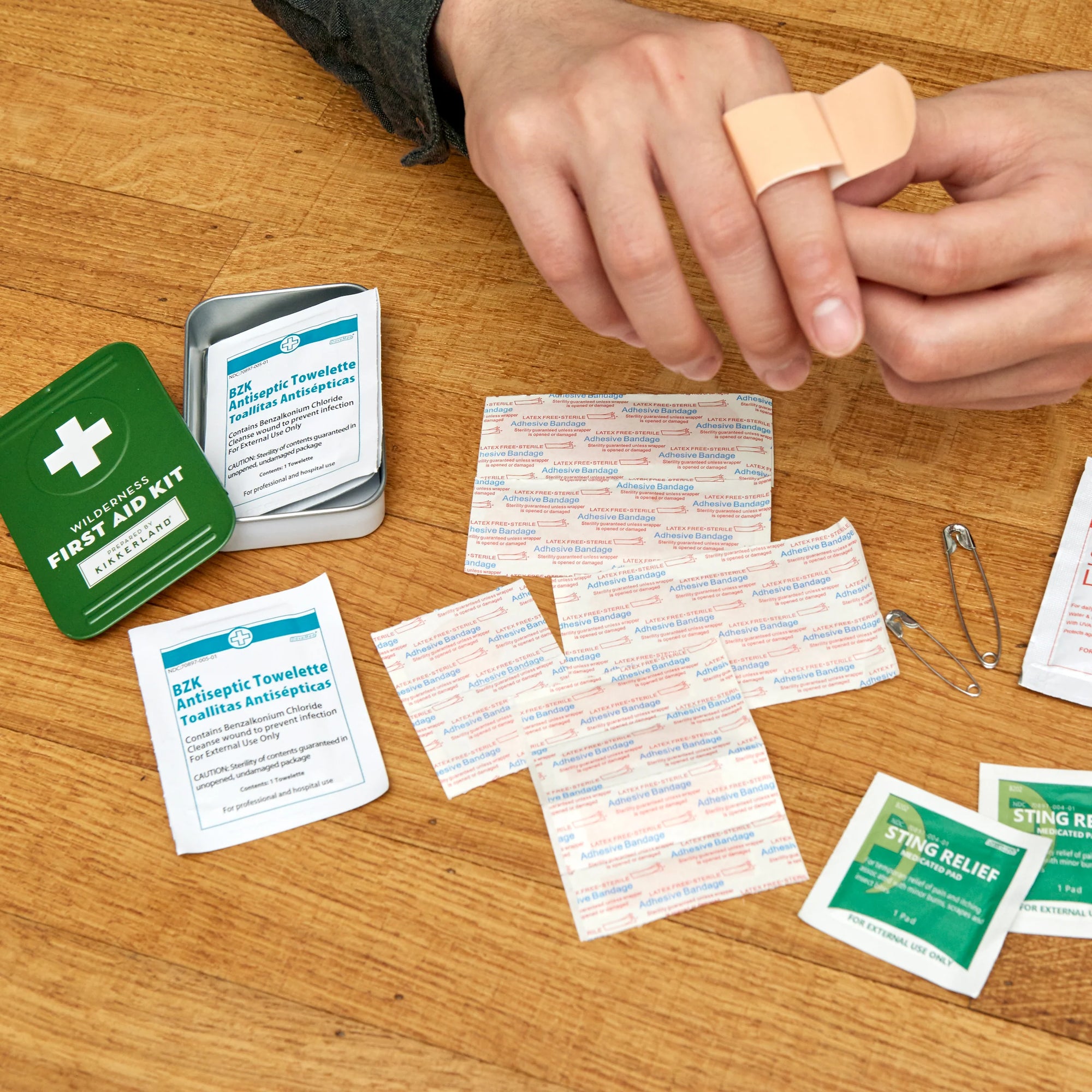 Person applying a bandage from a Wilderness First Aid Kit housed in durable metal casing, spread out on a wooden table, including antiseptic towelettes and sting relief pads.