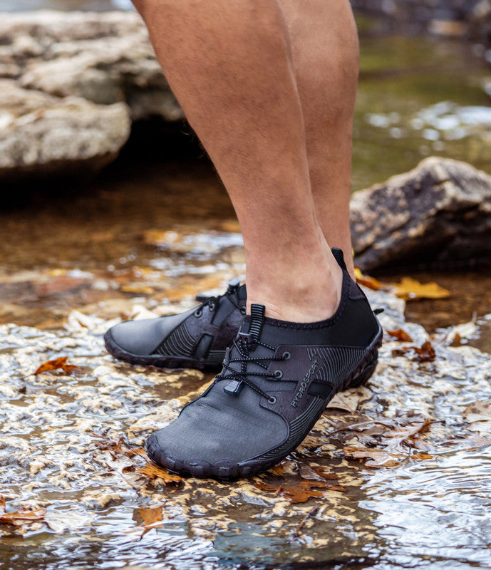 A person wearing HydroSport FitKicks with drainage holes stands in a shallow stream with leaves and rocks around.