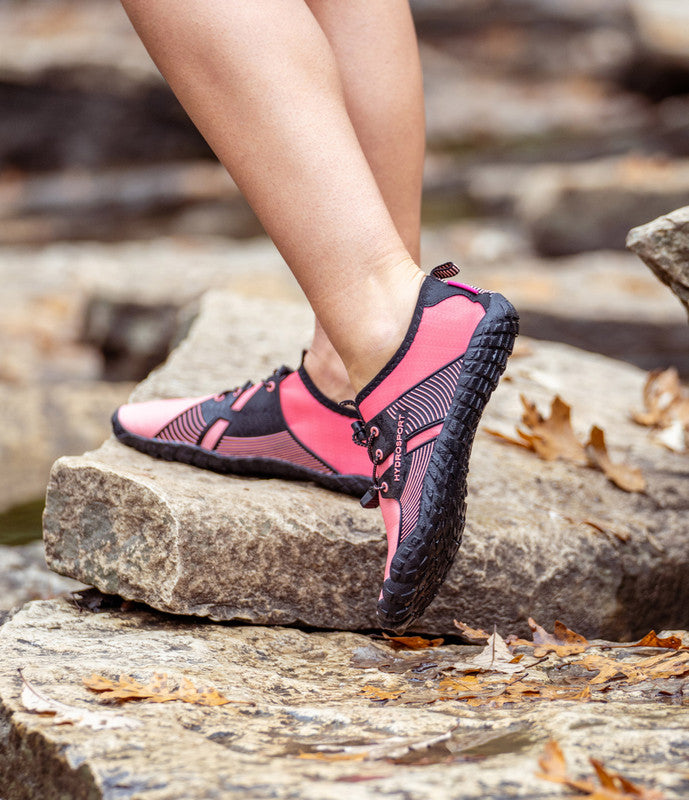 Close-up of a person's feet wearing pink and black HydroSport FitKicks, stepping on a rocky surface with scattered dry leaves.