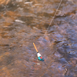 A Huckleberry Fishing Kit floats in a clear, rippling stream, attached to a fishing line near an outdoor popcorn cooking setup, indicating someone is enjoying both fishing and making popcorn nearby.