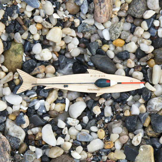 A Huckleberry Fishing Kit-shaped object lies on a pebble surface, marked with measurements and a red stripe, resembling a campfire popcorn popper.