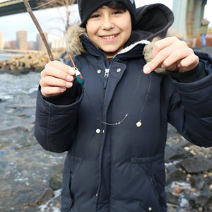 A young boy smiling at the camera, proudly holding up a Huckleberry Fishing Kit with two small fish, standing by a river near a campfire popcorn popper.