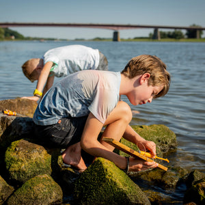 Two boys exploring a riverbank; one measuring an object with a Huckleberry Make Your Own Motor Boat on a rock, another in the background.