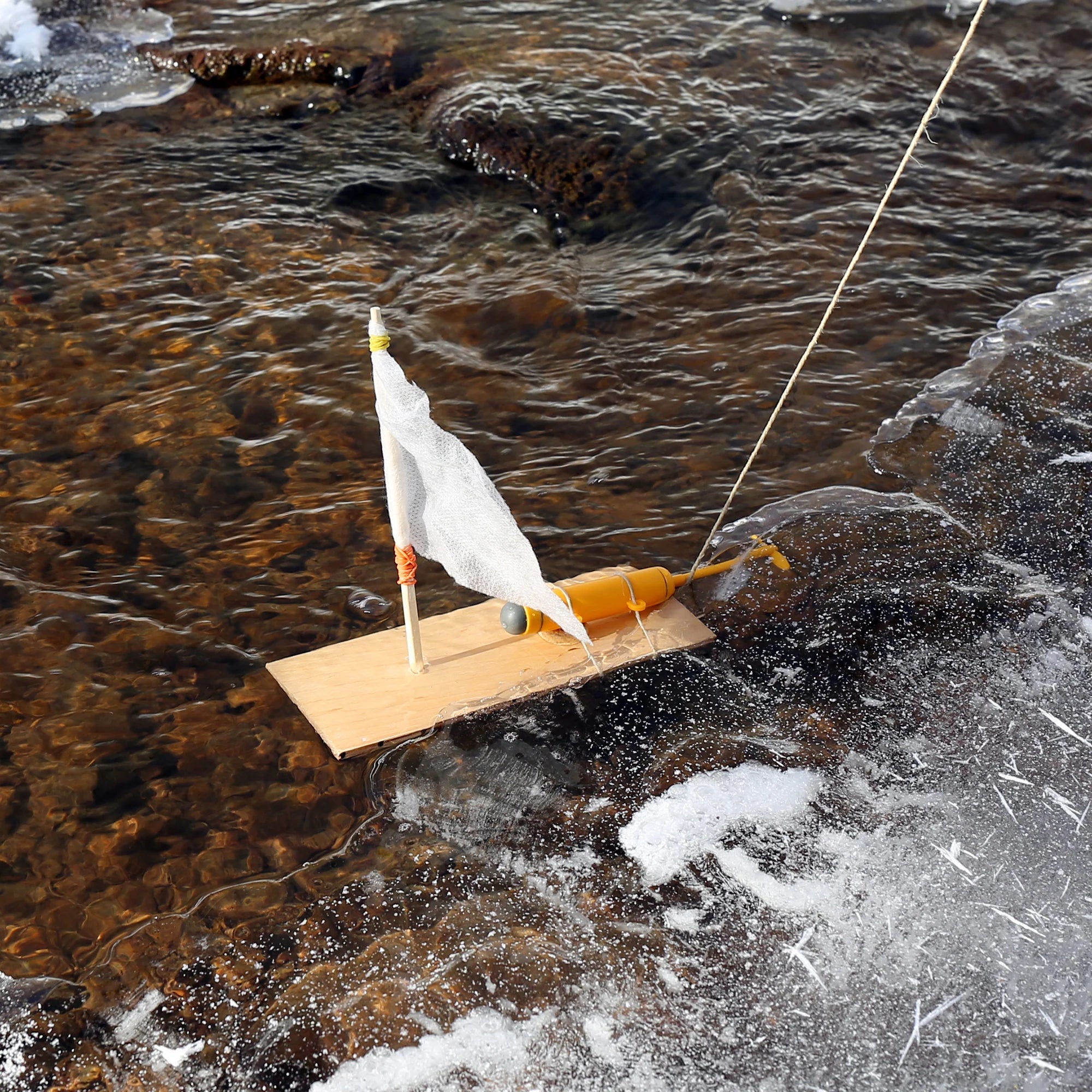 A Huckleberry Make Your Own Motor Boat with a white sail floats on a partially frozen stream, tethered by a string to the icy bank, perfect for any outdoors enthusiast.