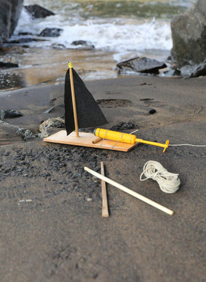 A small wooden toy Huckleberry Make Your Own Motor Boat on a sandy beach, with a fallen black sail, next to it a yellow raft, string, and wooden sticks from a sailboat building kit, with waves in the
