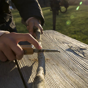 Person whittling a basswood stick with a Huckleberry Pocket Knife on a wooden table outdoors in sunlight.