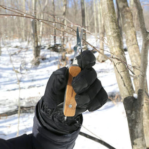 A person wearing black gloves holds a Huckleberry Pocket Knife near a basswood tree branch in a snowy forest setting.