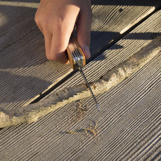 A hand holding a Huckleberry Pocket Knife to remove old varnish from basswood decking in sunlight.