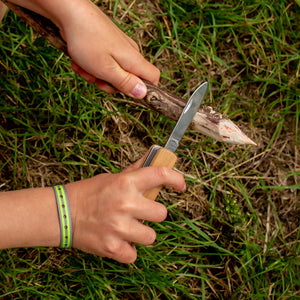Child's hands using a Huckleberry Pocket Knife to whittle a piece of basswood, with grassy background.